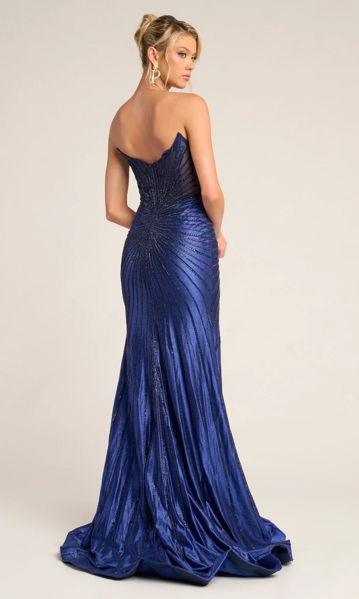 A woman in the Portia and Scarlett PS26250 long formal dress, a strapless, fitted blue beaded gown with a sunburst pattern, stands facing away and glances over her shoulder.